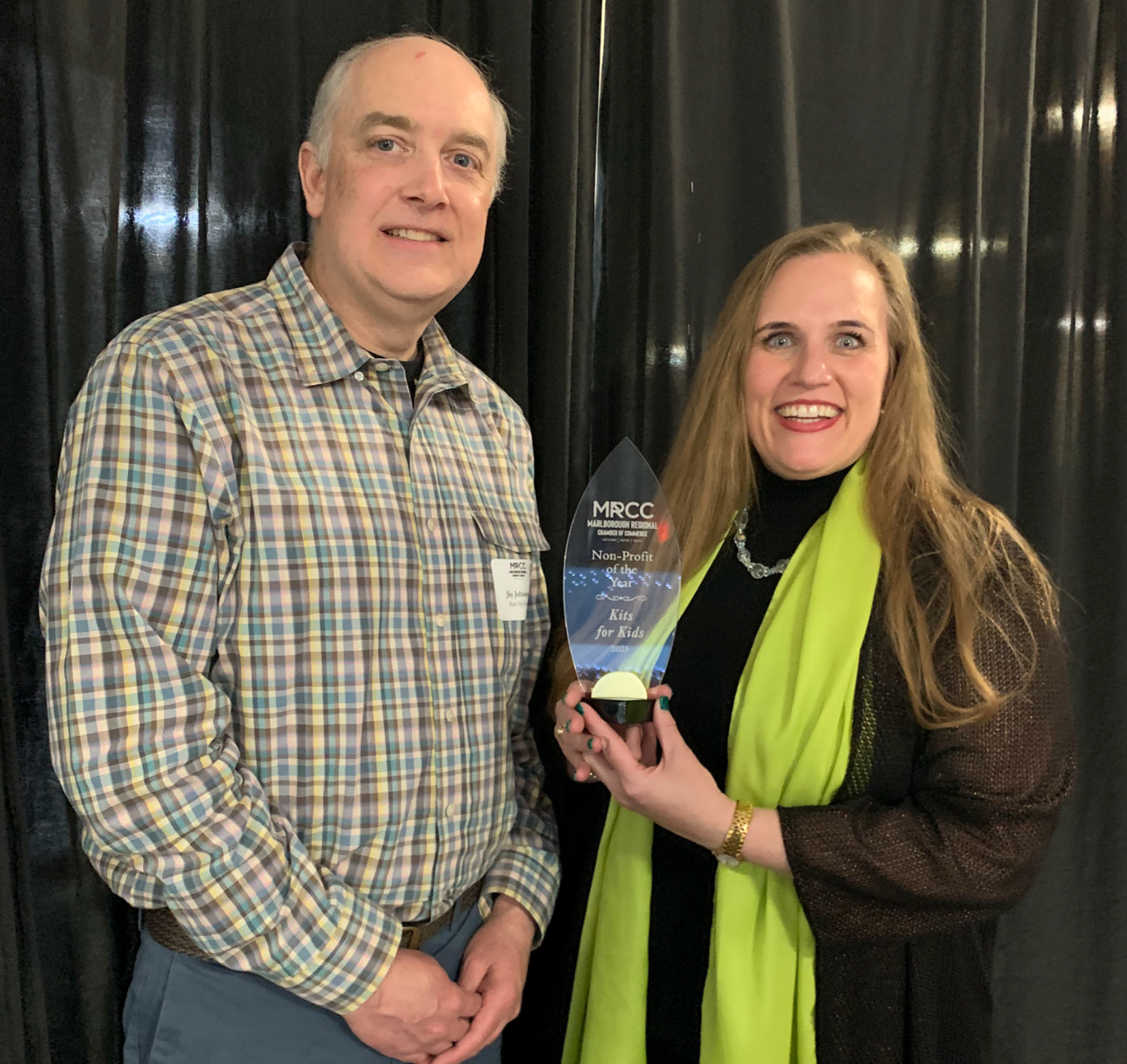 A man in a plaid shirt and a woman in a green scarf hold an award