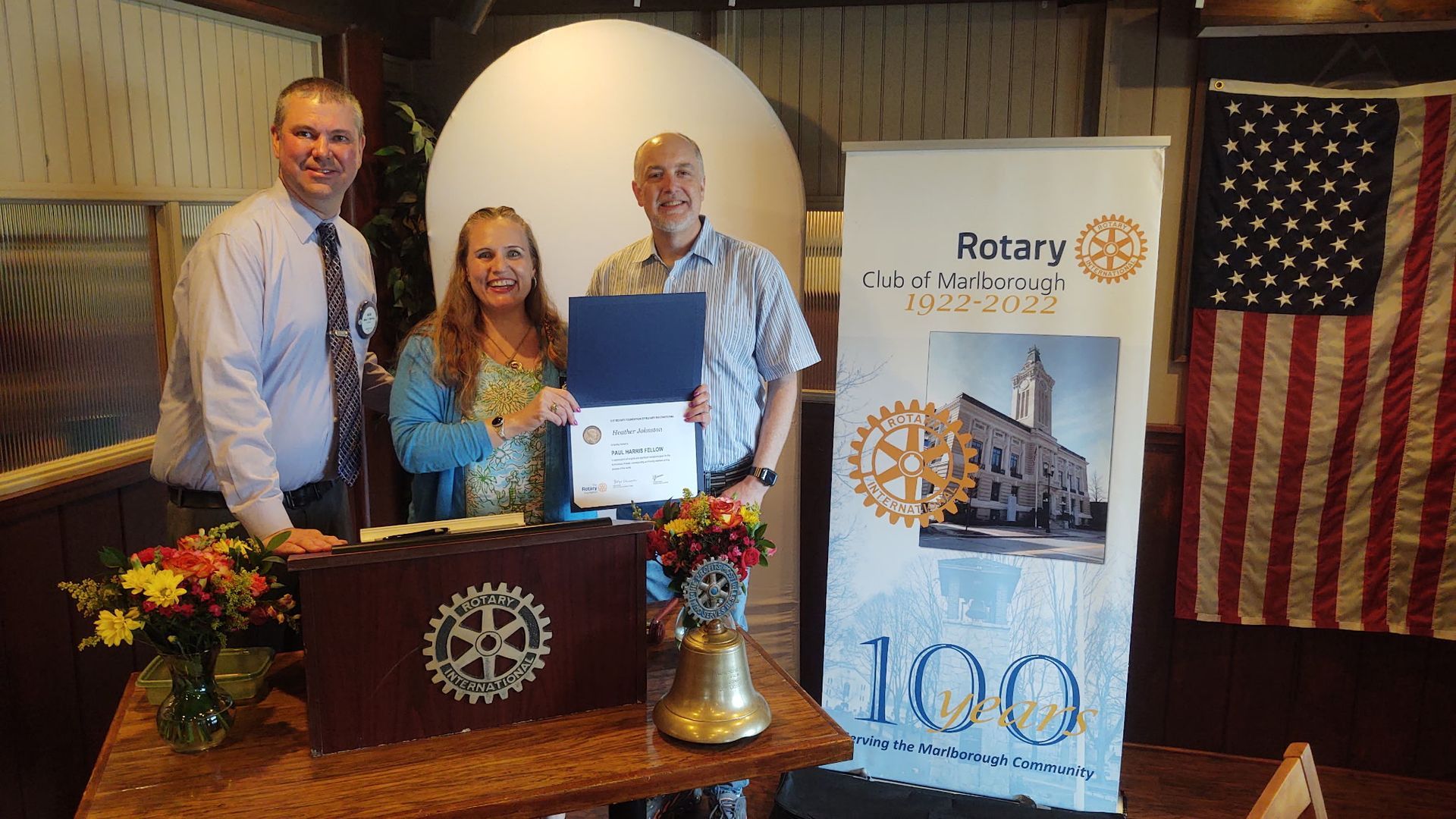 Three people at a Rotary event holding a certificate beside a banner and U.S. flag