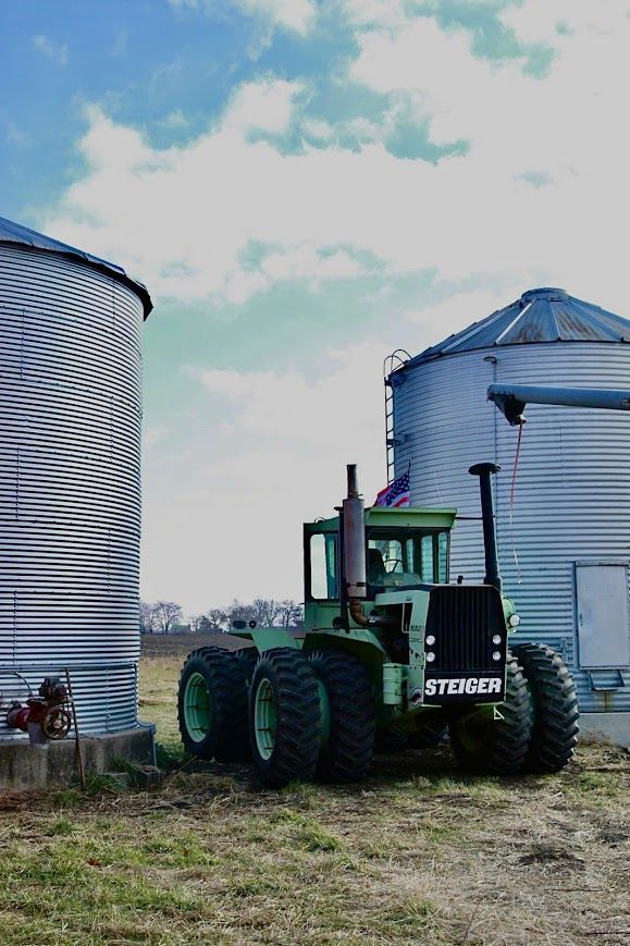 A green tractor is parked in a field next to two silos.
