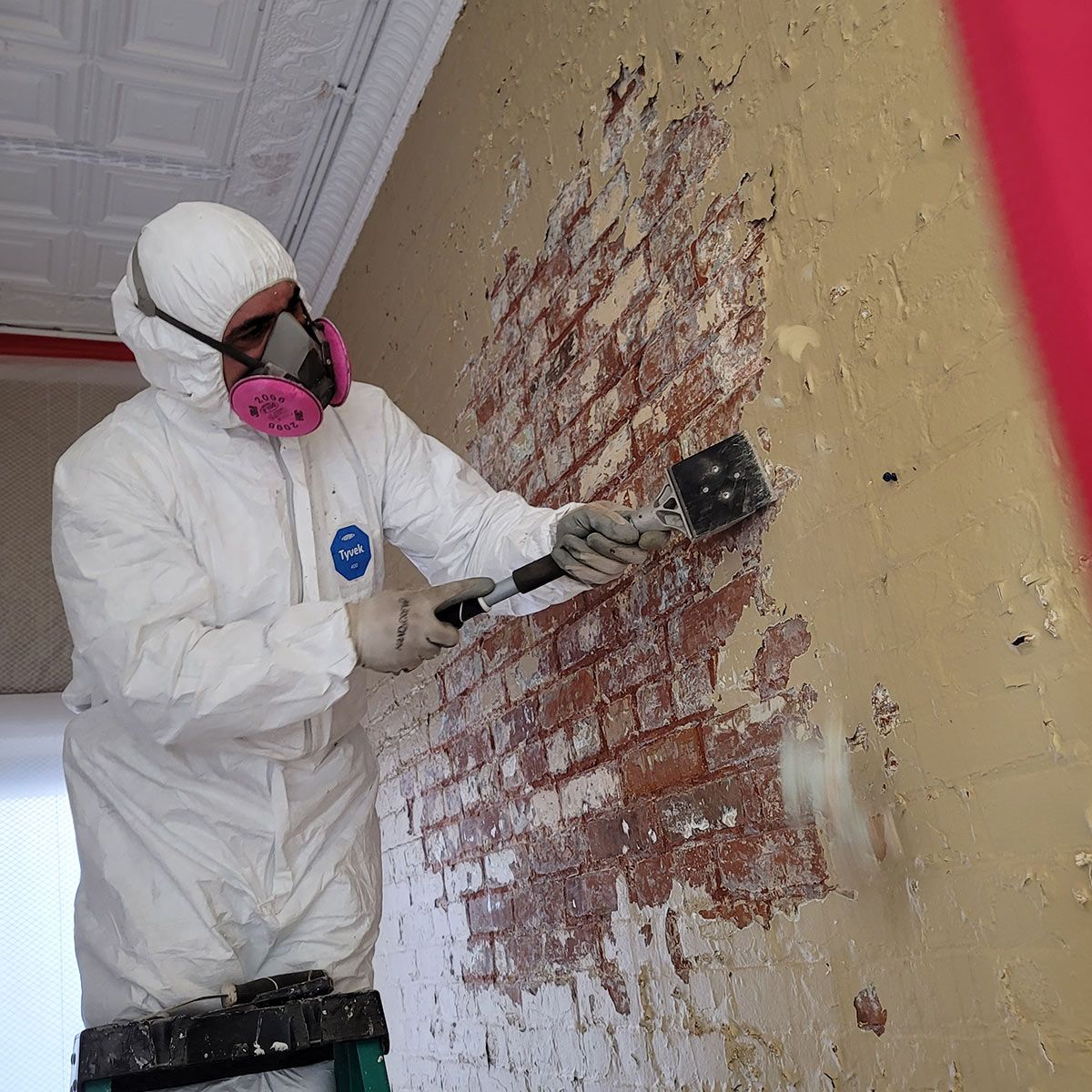 A man in a protective suit is cleaning off lead paint with a spatula.