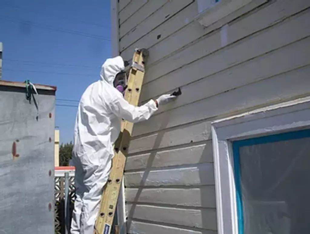 A man in a protective suit is painting over Lead Paint on the side of a house.