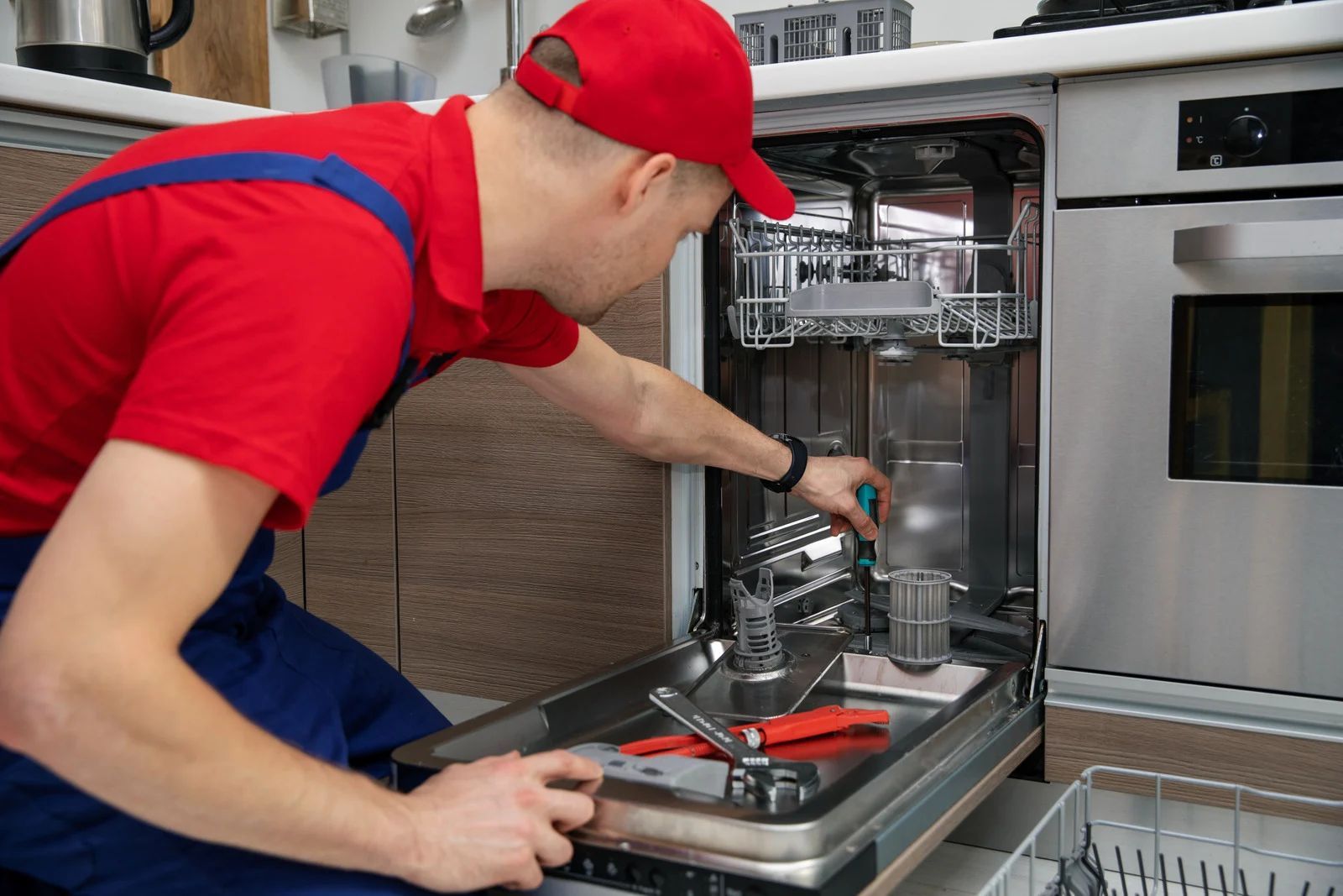 Man in red shirt and cap repairing a dishwasher in a kitchen.