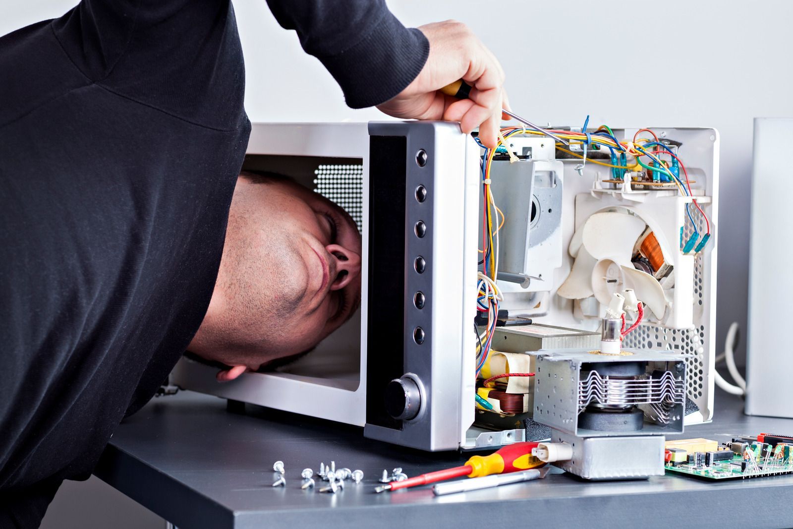 Person repairs microwave, head inside. Wires, components exposed. Screwdriver visible on table.