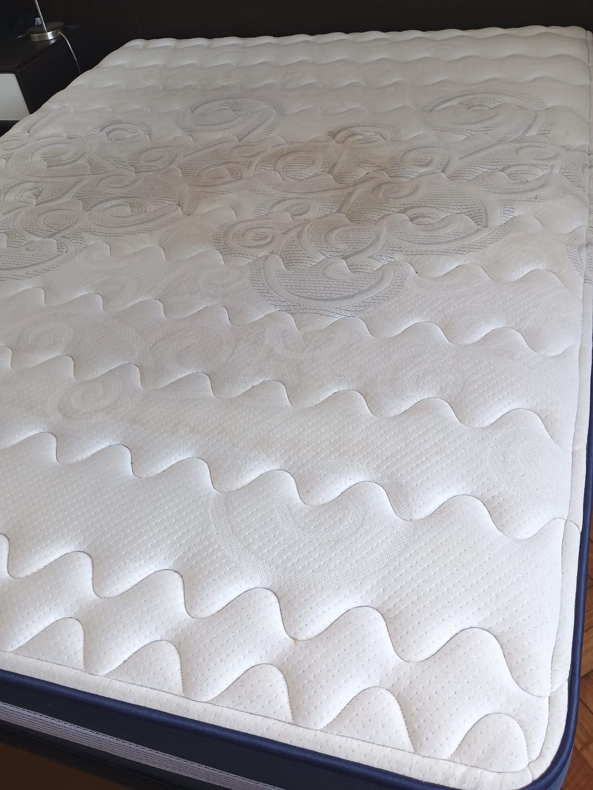Un colchón con una superficie blanca acolchada que presenta un sutil estampado floral gris, ribeteado con tela azul oscuro.