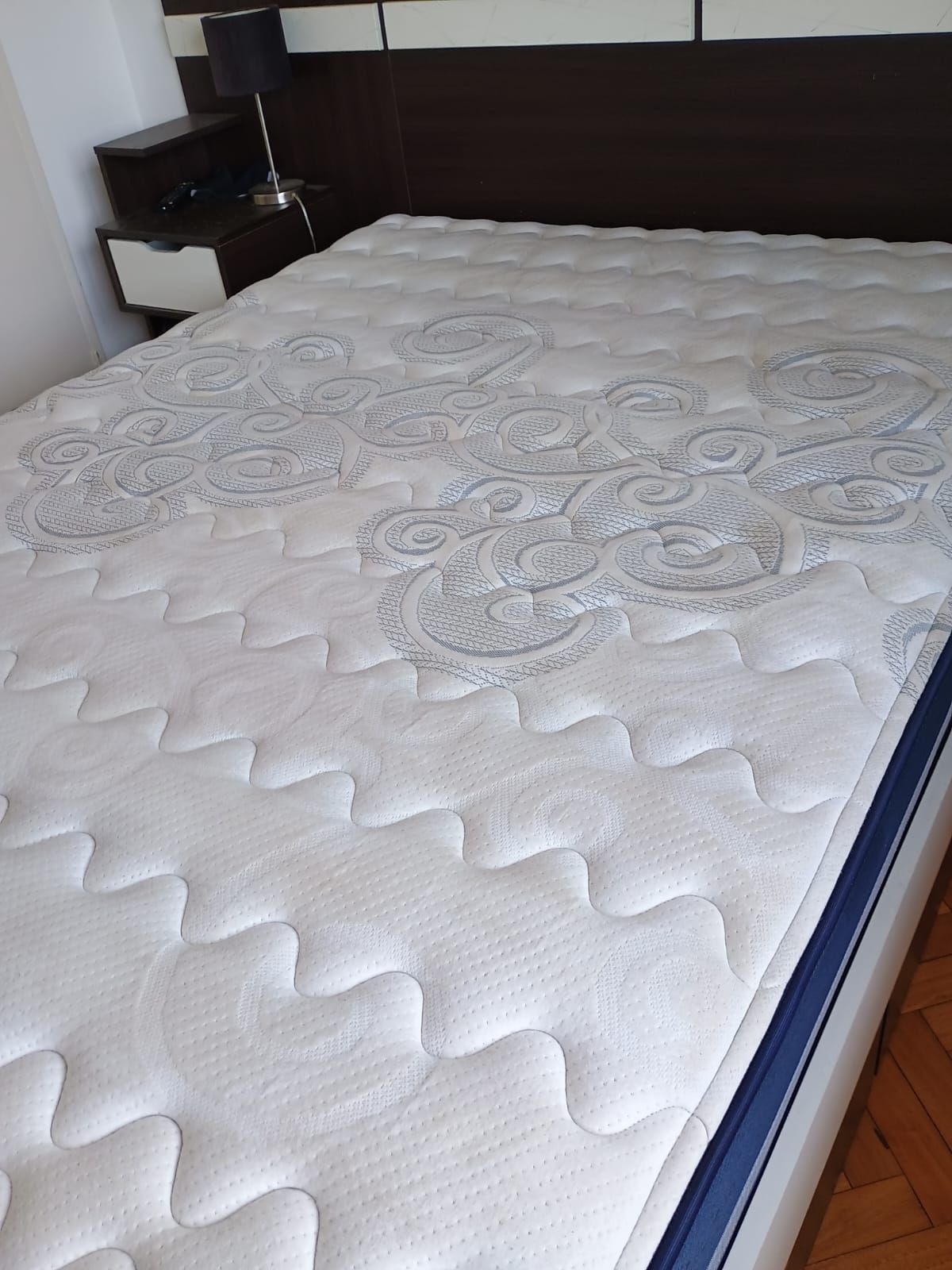 Un colchón blanco con un estampado de volutas grises, colocado frente a un cabecero marrón oscuro en un dormitorio.