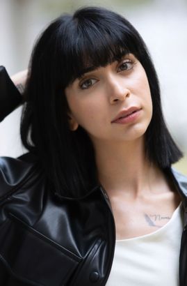 Woman with dark hair and bangs, wearing a black leather jacket, looking at the camera.