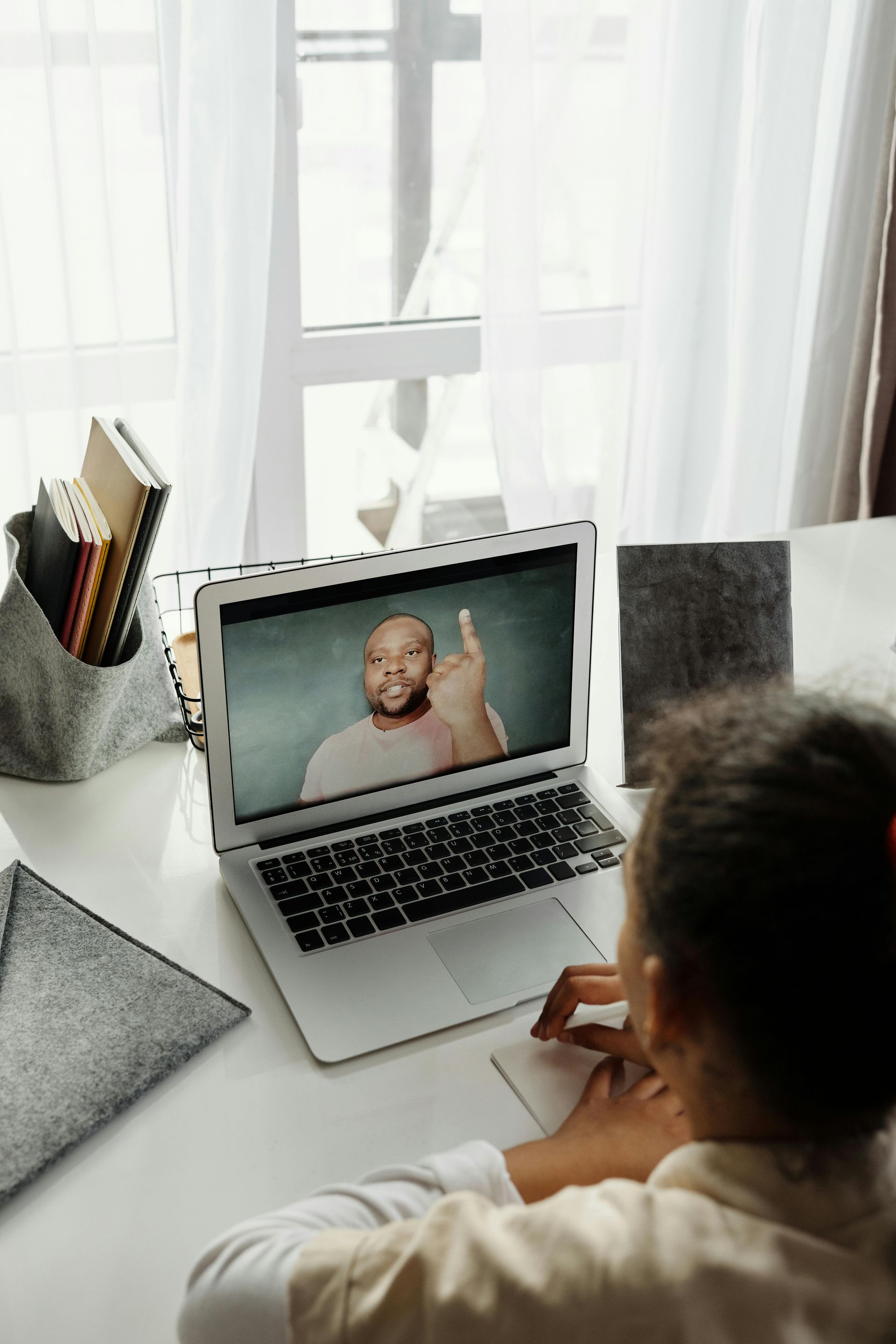 Girl in video call with teacher on laptop, writing at desk.
