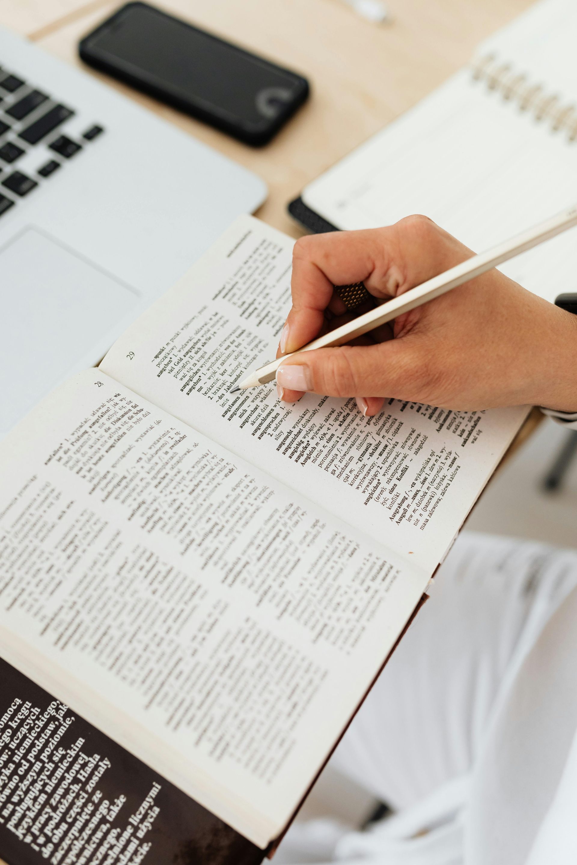 Person writing in a book with a pencil, near a laptop, phone and notepad on a desk.