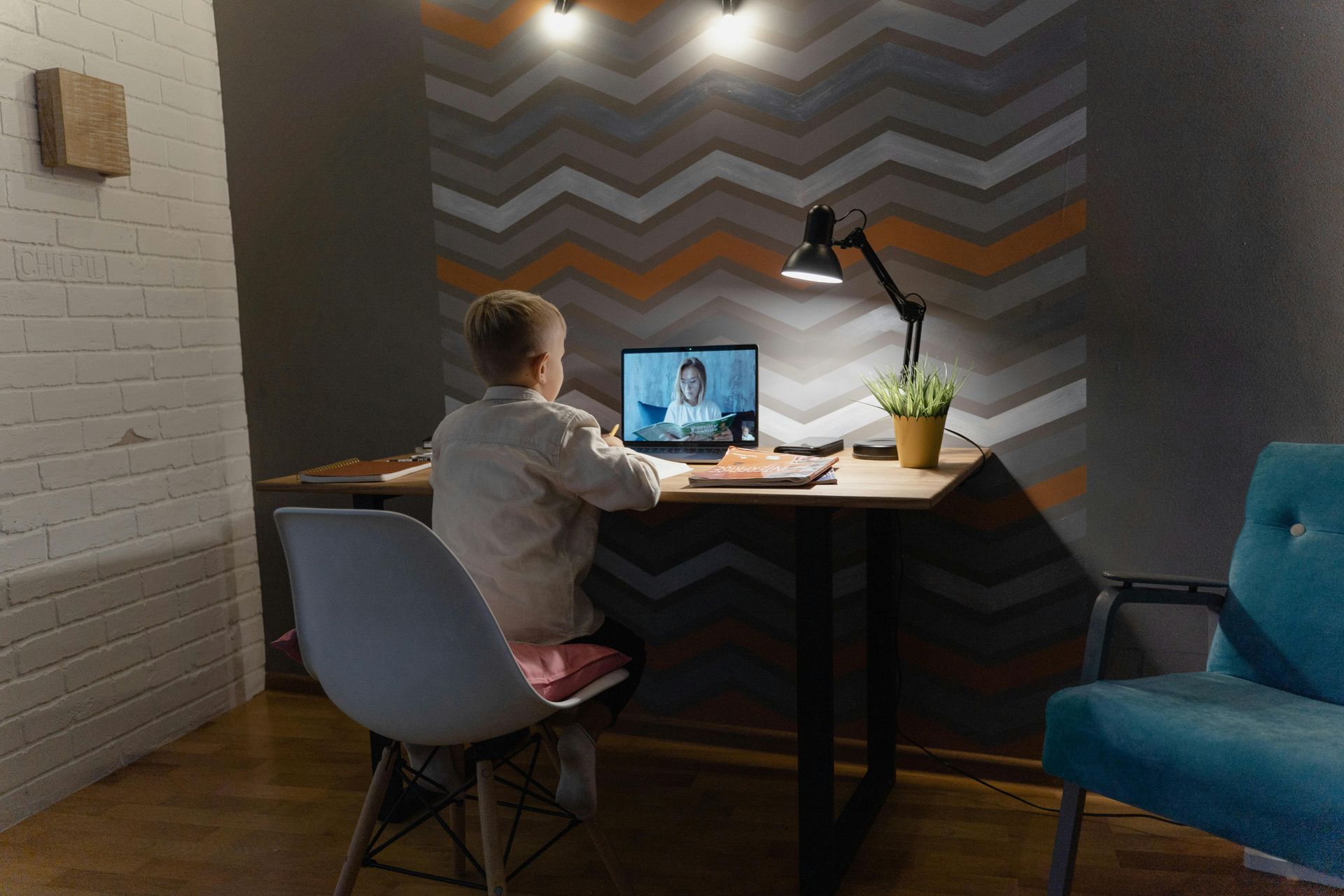 Boy at desk, video-conferencing on laptop, with lamp, plant, and gray chevron wall.