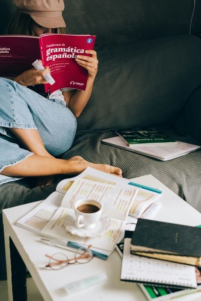 Woman studying on a couch, reading a book with a coffee, books, and notes on a table.