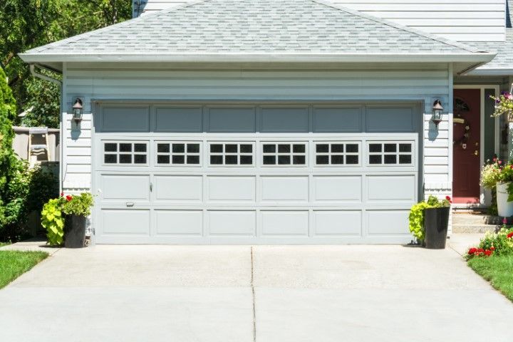 Gray garage door with windowed top panels on a house. Concrete driveway, green lawn, flower pots.
