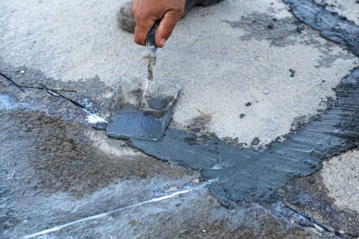 Hand using a trowel to apply dark sealant to cracks in a light gray concrete surface.