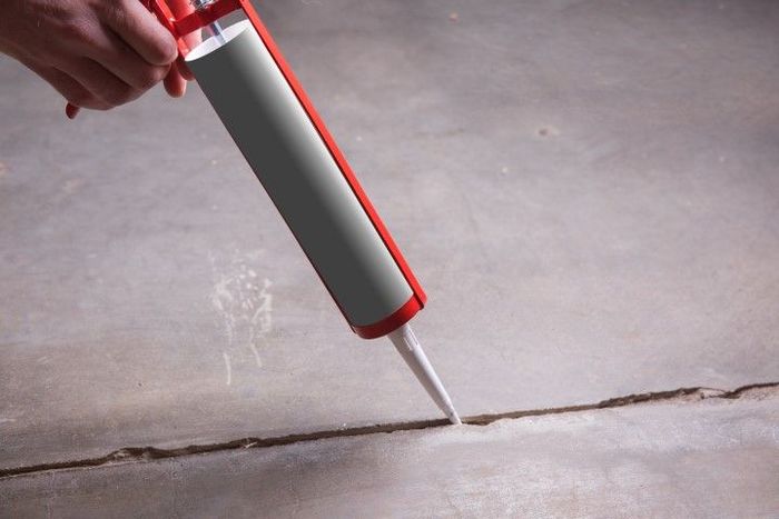 Hand holding caulking gun, applying sealant to a crack in a concrete floor.