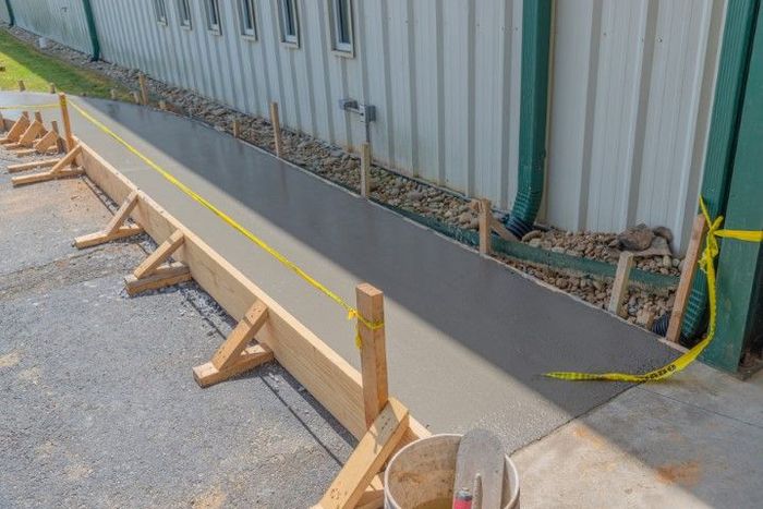 Freshly poured concrete sidewalk next to a building, yellow tape, wooden supports.