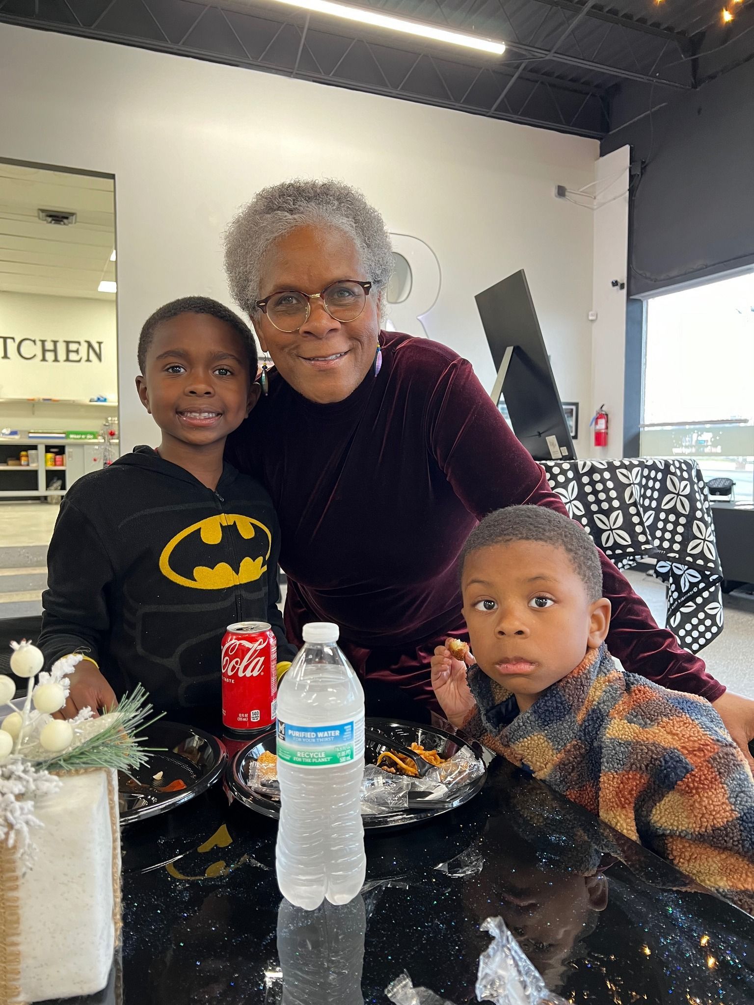 A woman is sitting at a table with two children . one of the children is wearing a batman shirt.
