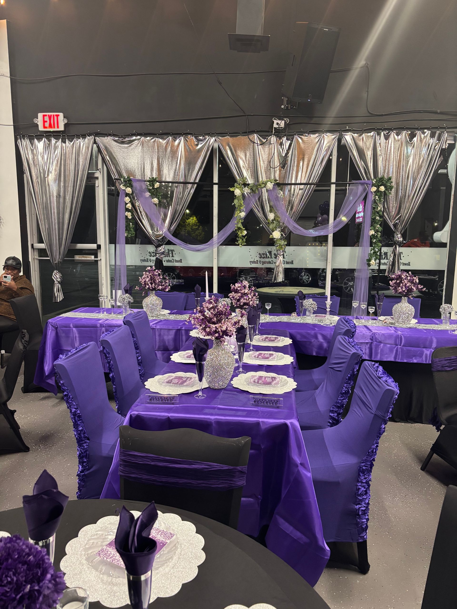 Tables set for an event, featuring purple tablecloths, chair covers, and centerpieces, in a room with large windows.