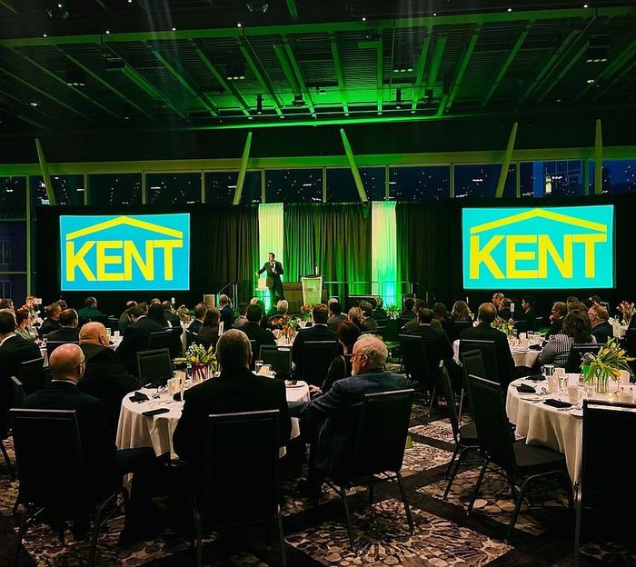 Audience at Kent event; speaker on stage; large screens with Kent logo; green lighting.
