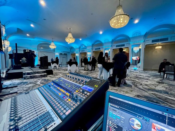 Sound mixing board in front of a blue-lit event space. Tables, people, stage, and chandeliers visible.