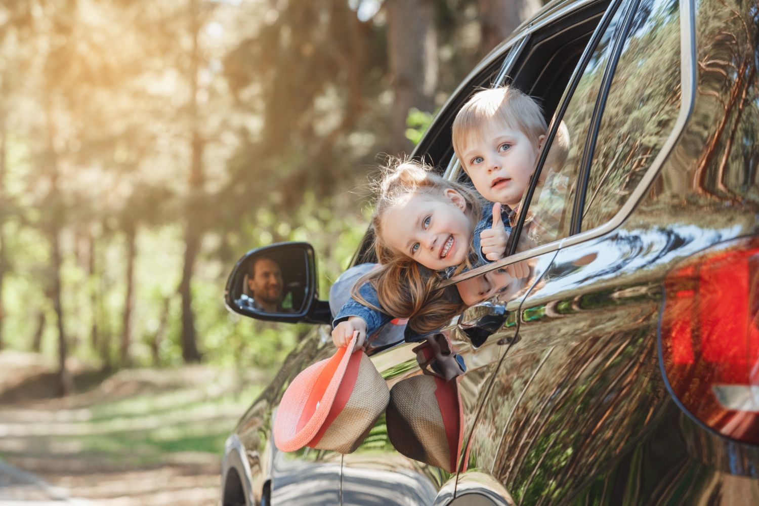 Two children leaning out of a car window on a sunny roadside, smiling with bags in hand