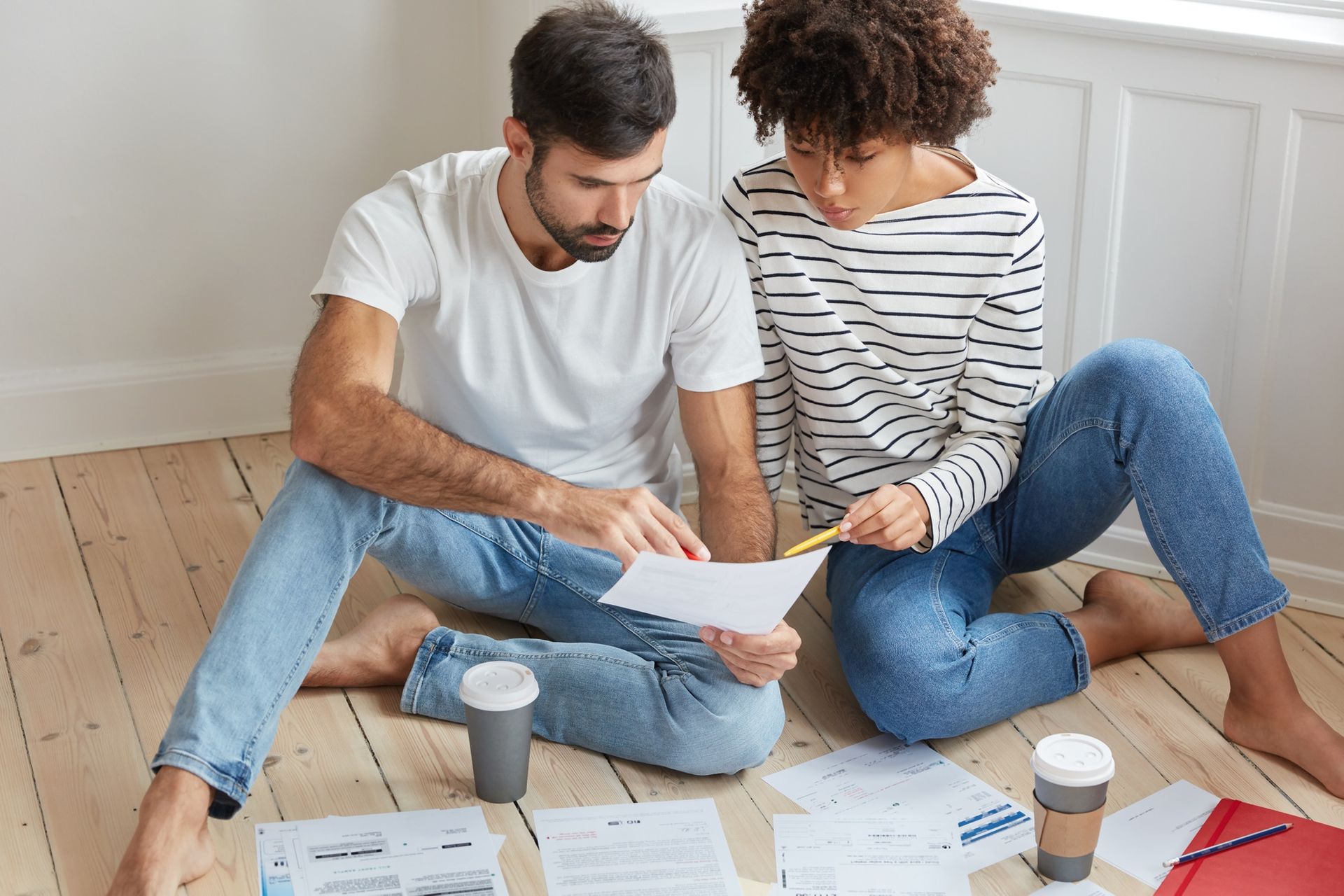 Two people sit on the floor reviewing papers and bills beside coffee cups in a bright room.