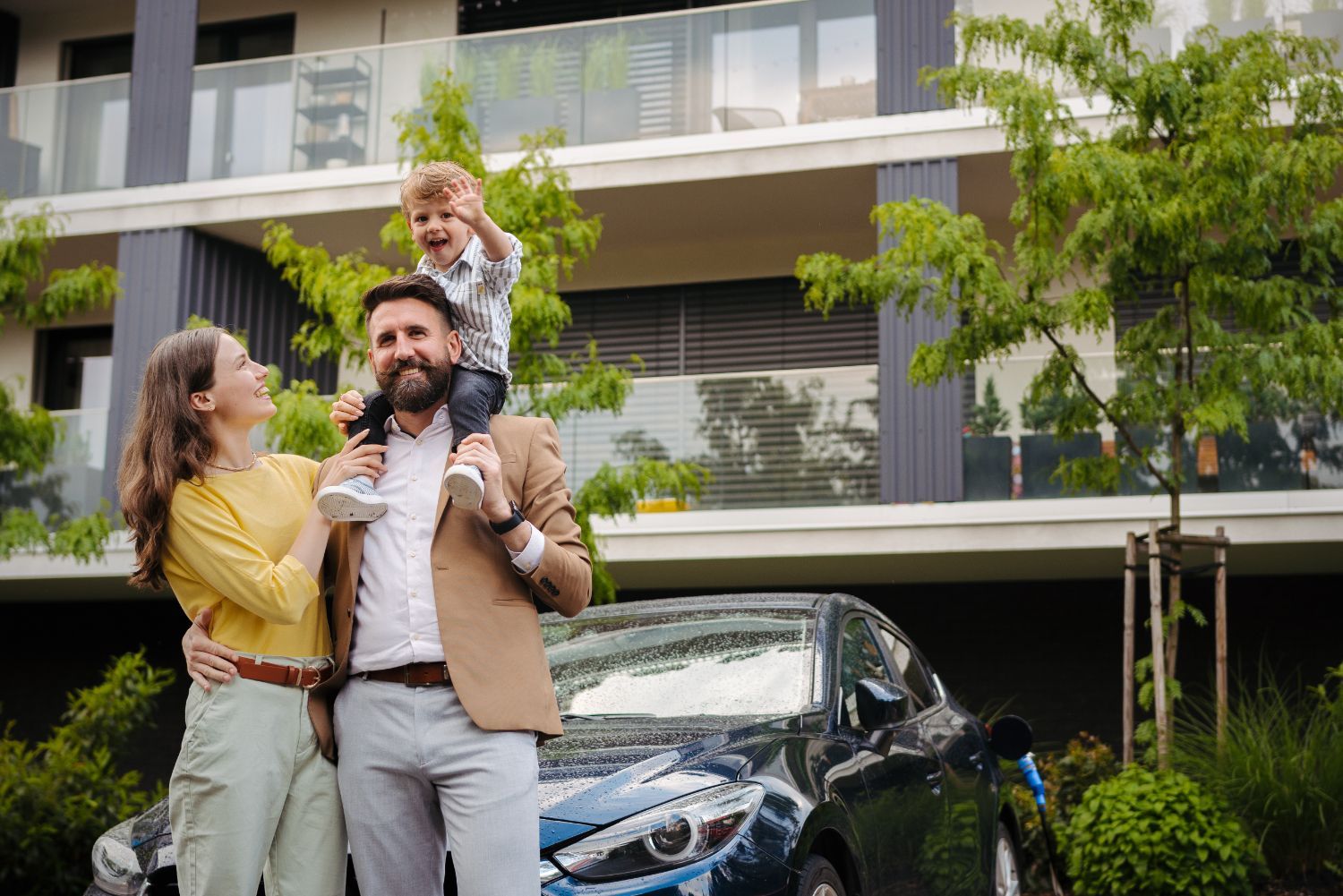 Family standing by black car outside modern house; child sits on father’s shoulders, smiling.