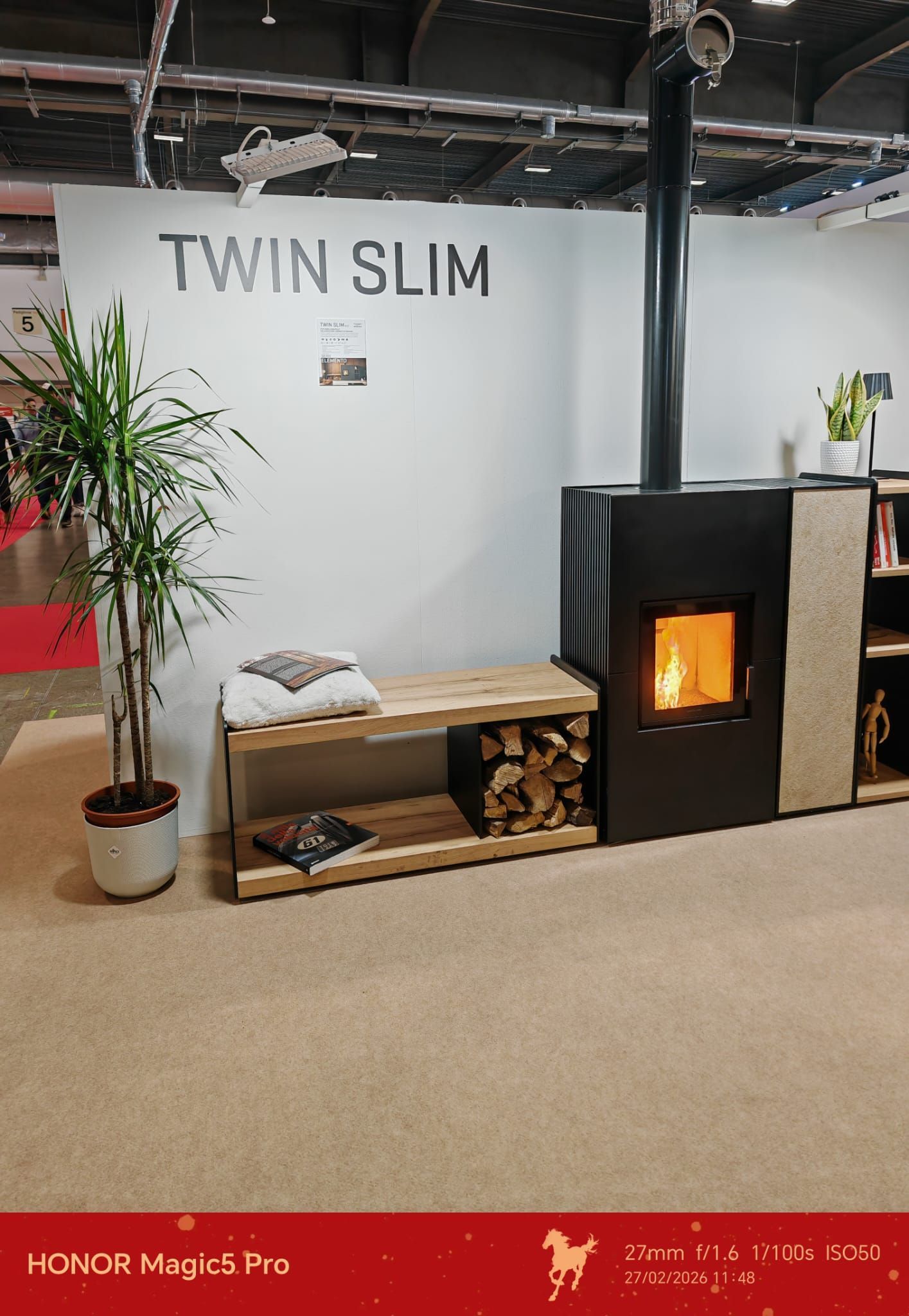 Camino con legnaia, panca e pianta alta contro una parete bianca con la scritta 