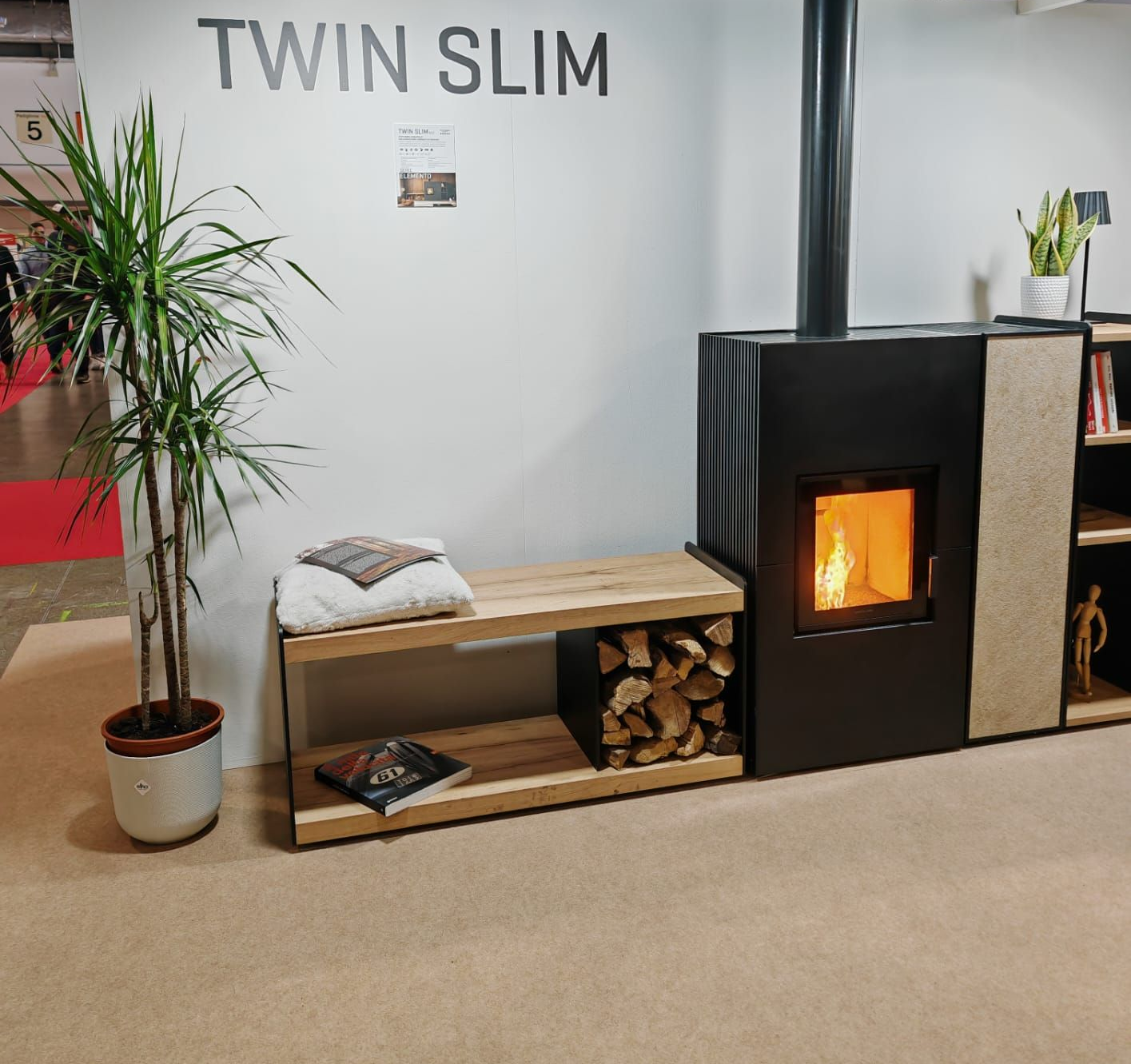 Camino moderno con legnaia e panca, affiancato da piante contro una parete bianca.