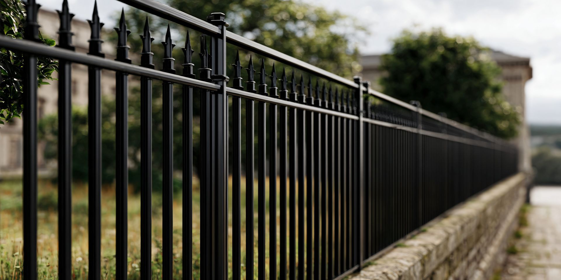 Black metal fence in front of a stone wall, leading to a blurred background of greenery and a building.