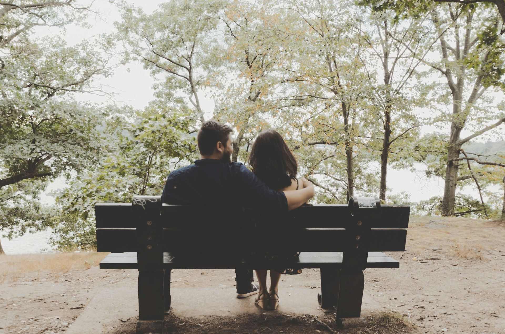 A man and a woman are sitting on a park bench looking out over a lake.