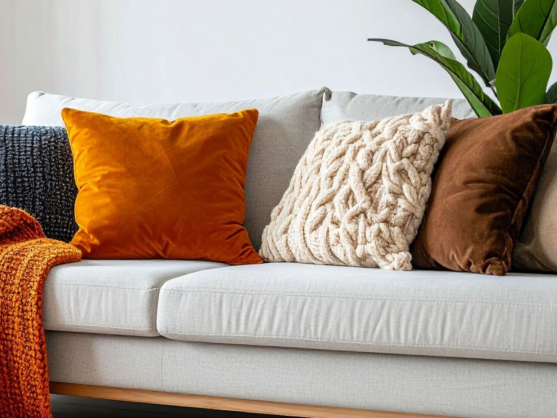 A light gray sofa decorated with a mix of solid velvet orange, brown, and textured cream pillows, with a plant nearby.