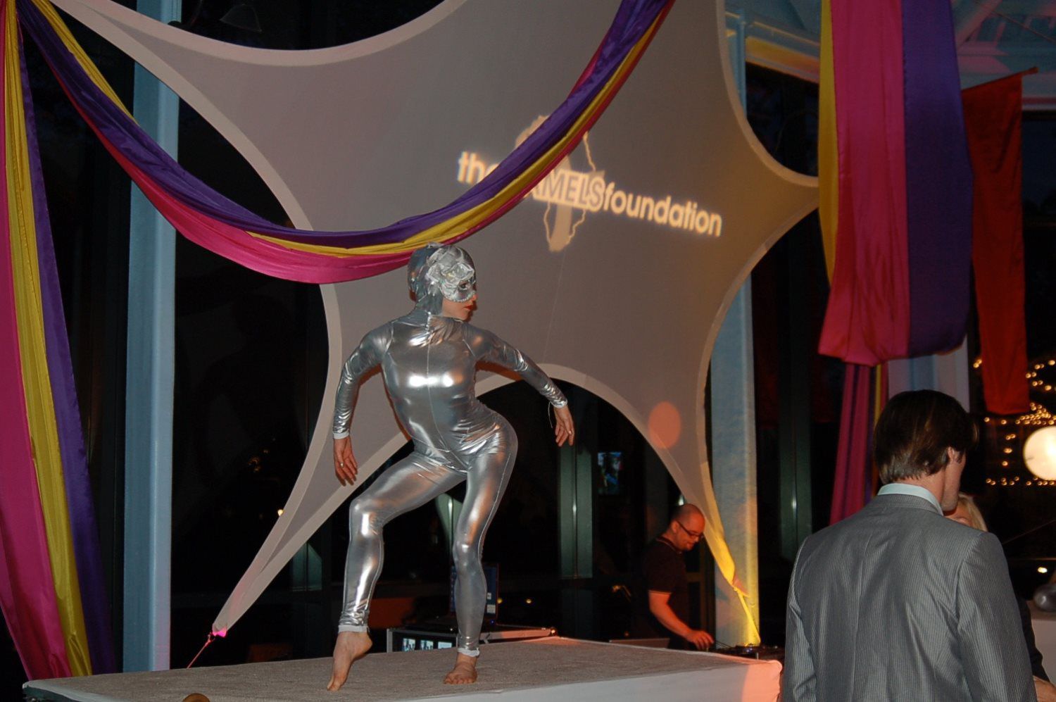 A woman in a silver bodysuit is dancing on a stage with the word foundation projected on the wall behind her