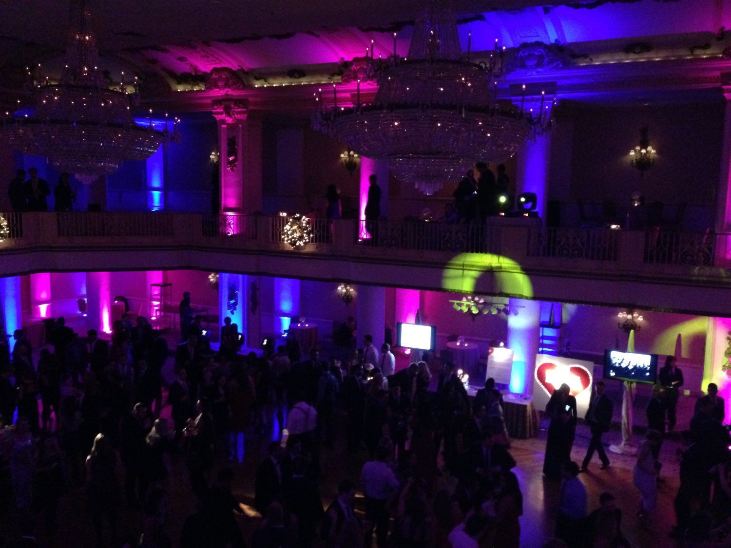 A large crowd of people are dancing in a ballroom with purple and green lights