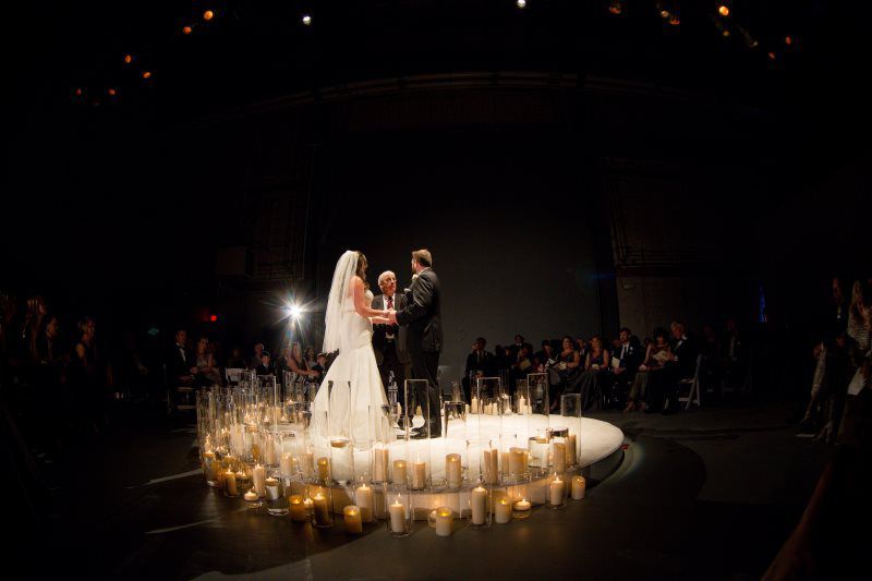 A bride and groom are standing on a stage surrounded by candles.