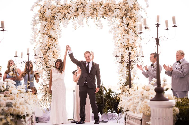 A bride and groom are holding hands at their wedding ceremony under a floral arch.