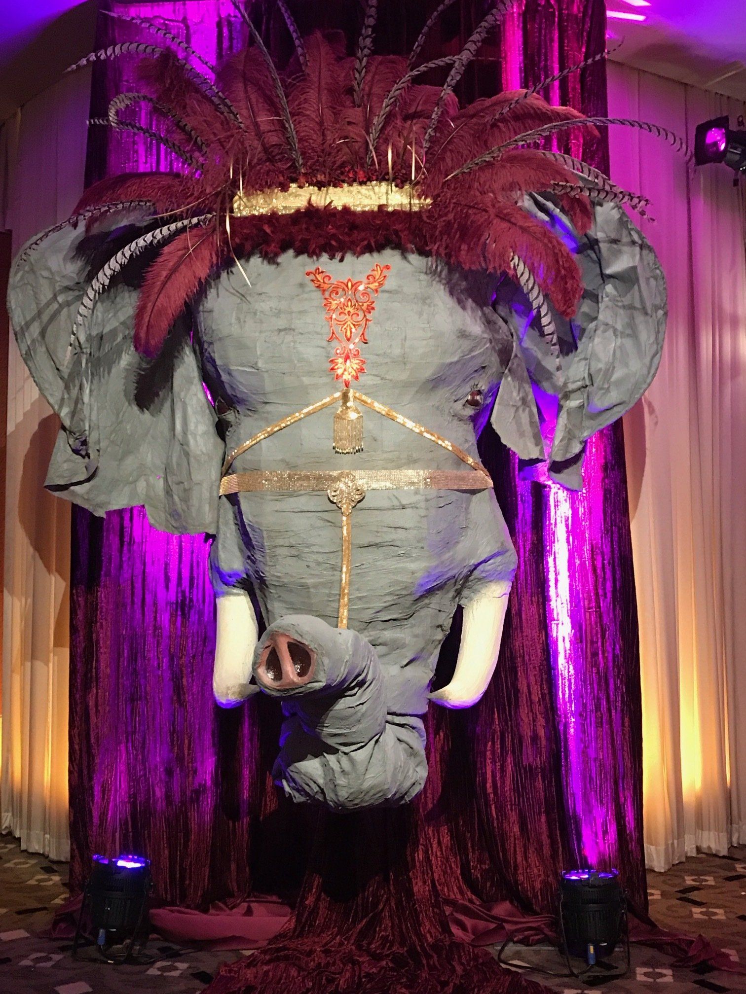 A large stuffed elephant with feathers on its head is hanging from a pole.