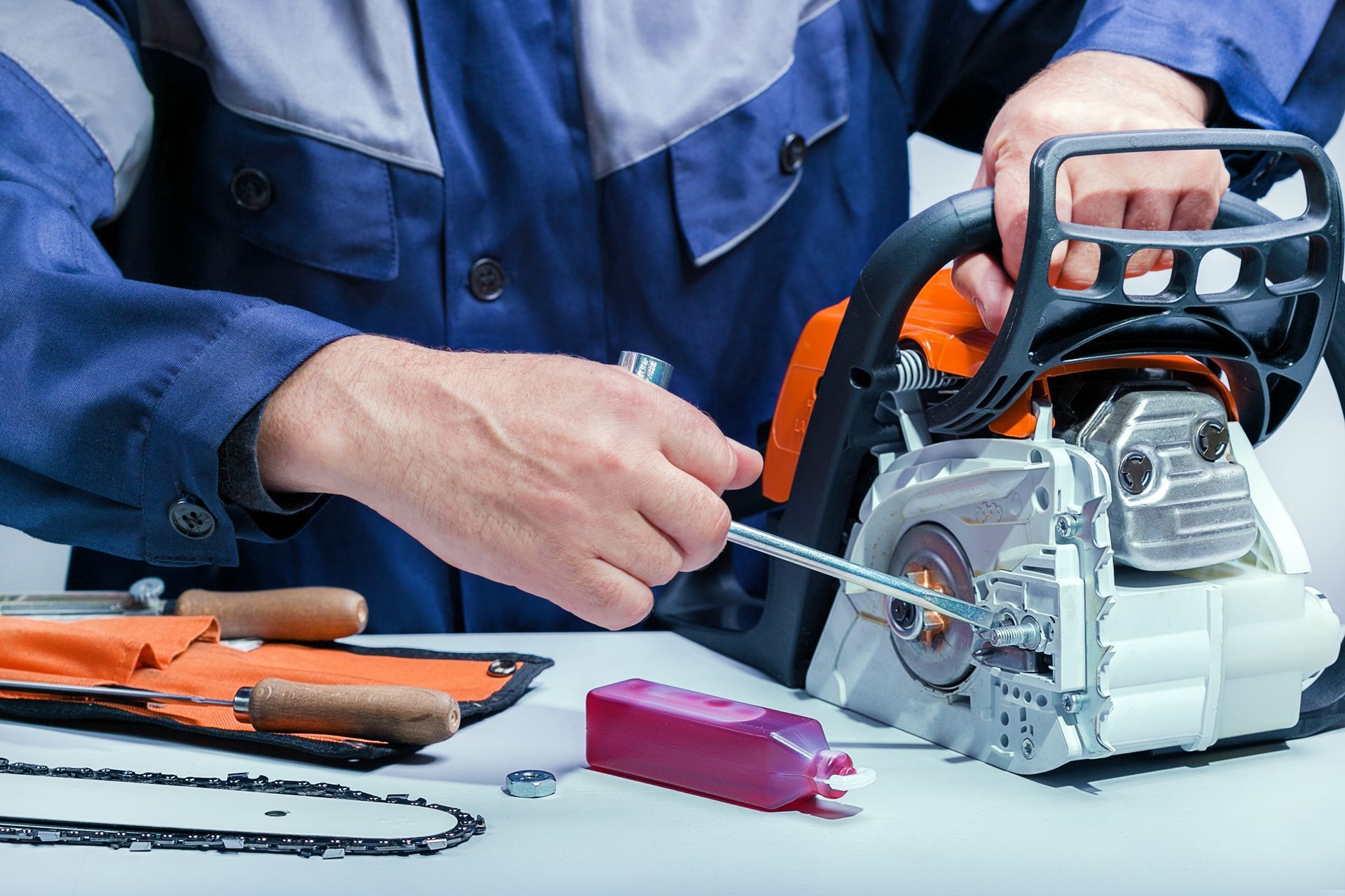 Person in blue overalls servicing a chainsaw, with tools and chain laid out.
