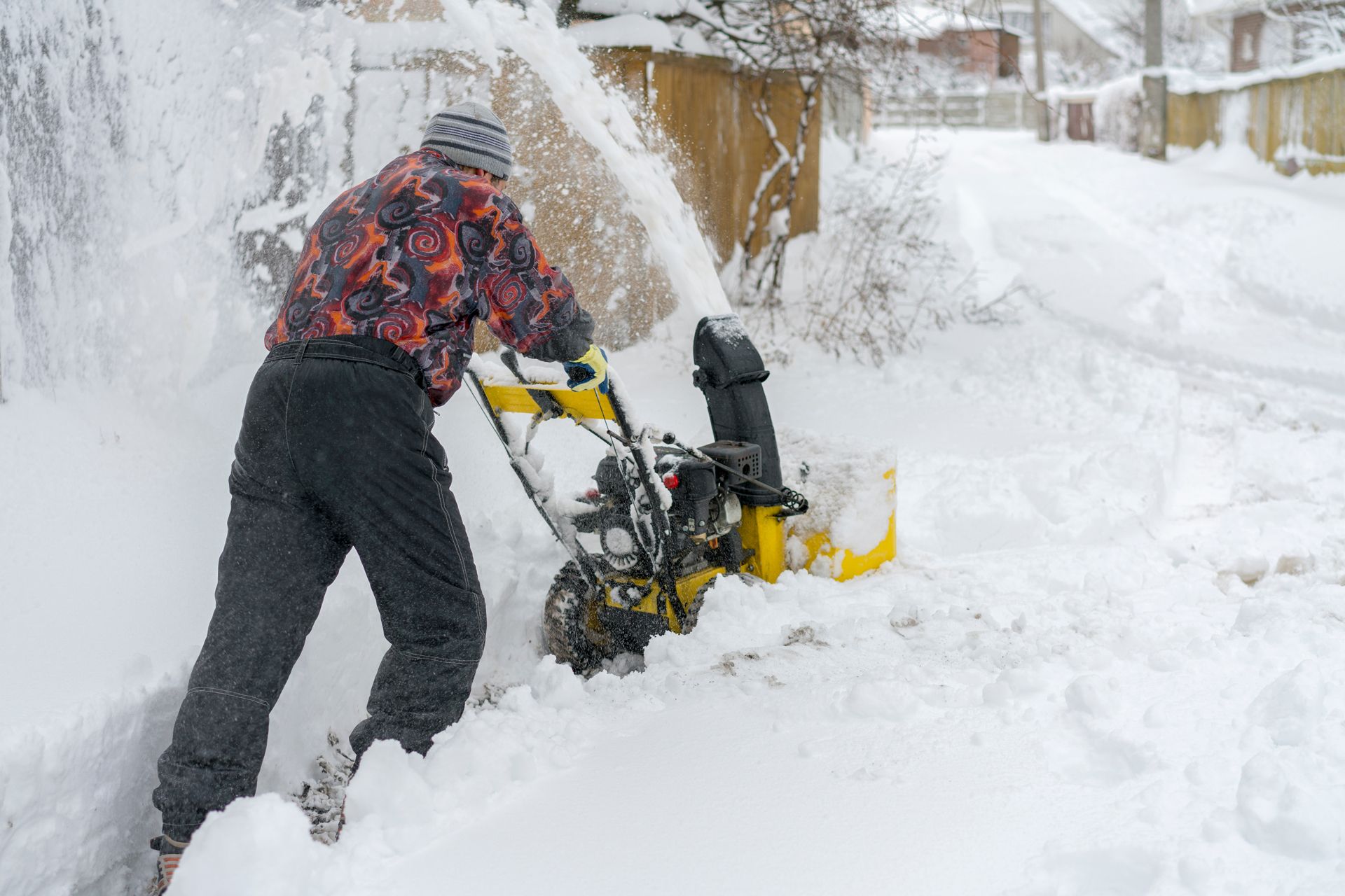 Man using a yellow snowblower to clear a snowy driveway on a winter day.
