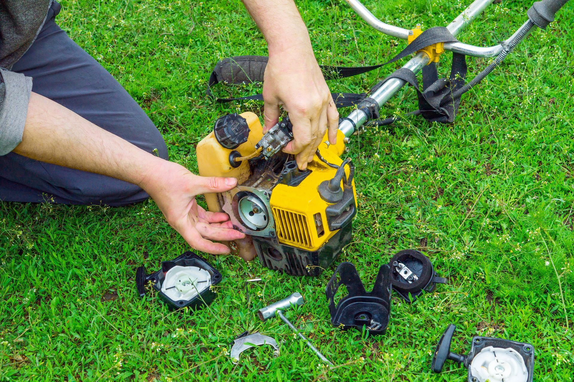 Person disassembling a yellow weed wacker on green grass, parts scattered.