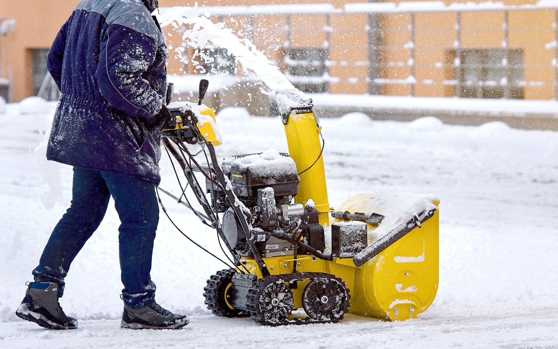 Person using a yellow snow blower to clear snow from a paved area in front of a building.