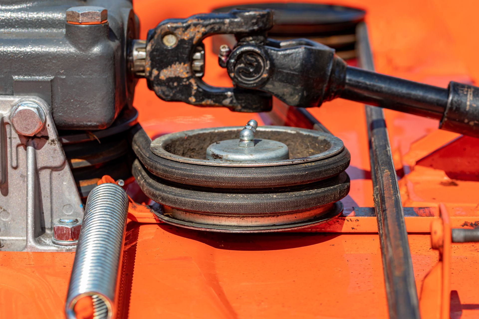 Close-up of a lawnmower deck showing a pulley, belt, and drive shaft on an orange surface.