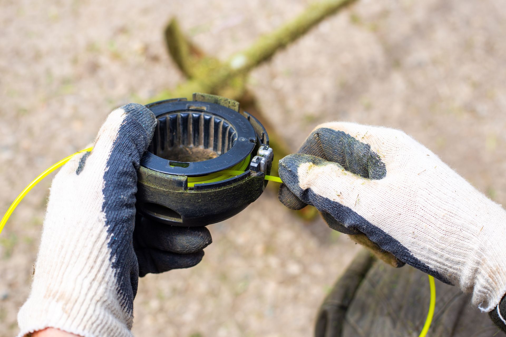 Gloved hands winding yellow trimmer line onto a black spool head, with a lawnmower in the background.