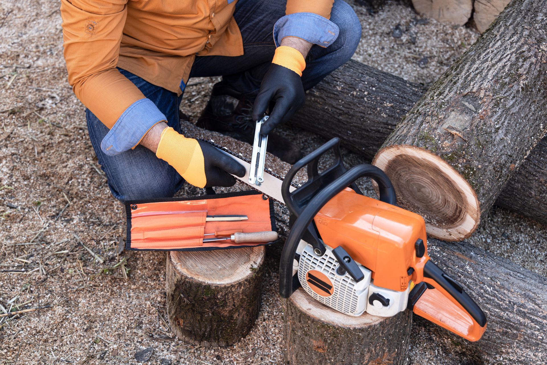 Person sharpening a chainsaw outdoors. Orange jacket, black gloves, tool kit, logs.