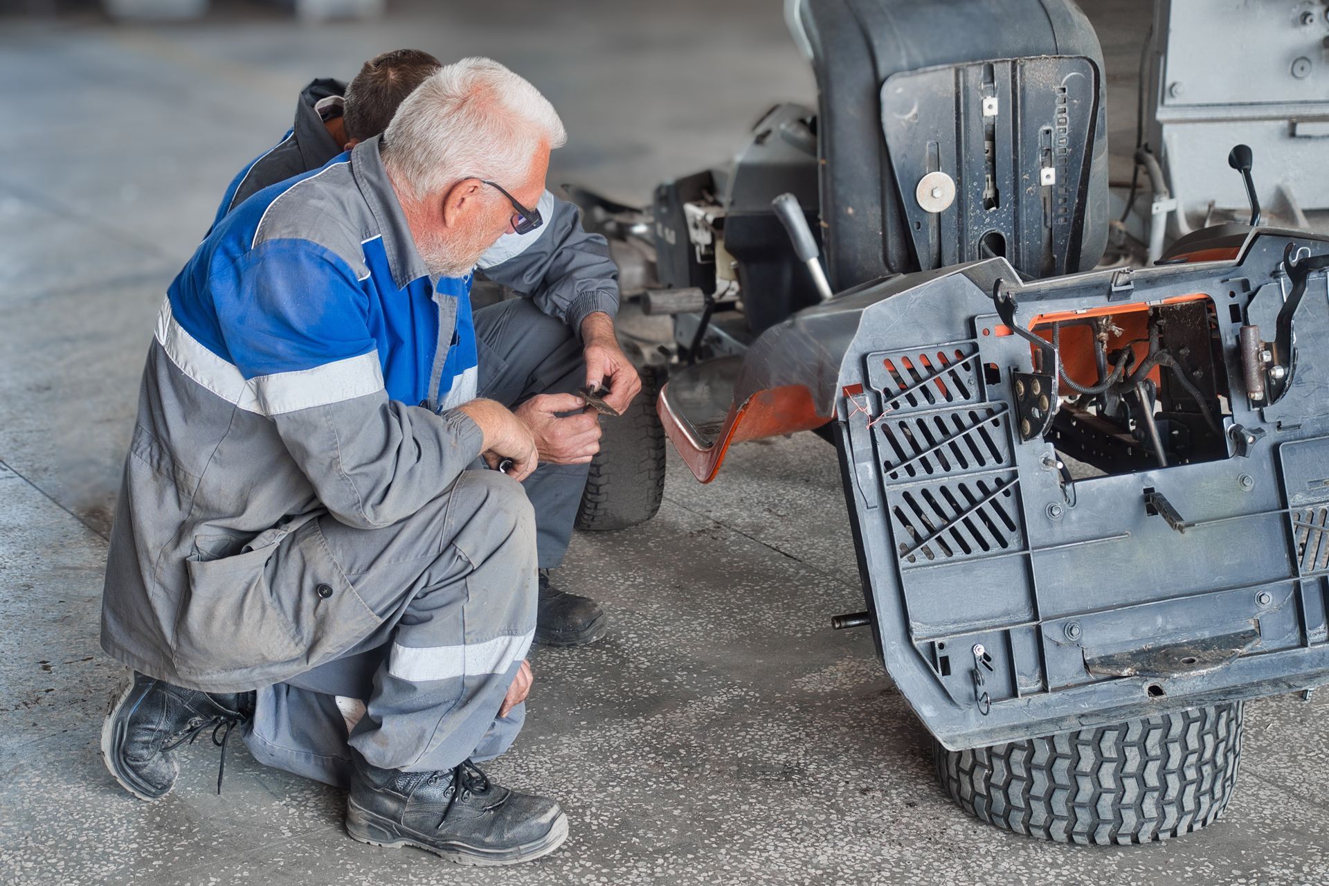 Man in blue and gray jumpsuit repairing a riding lawnmower in a workshop.