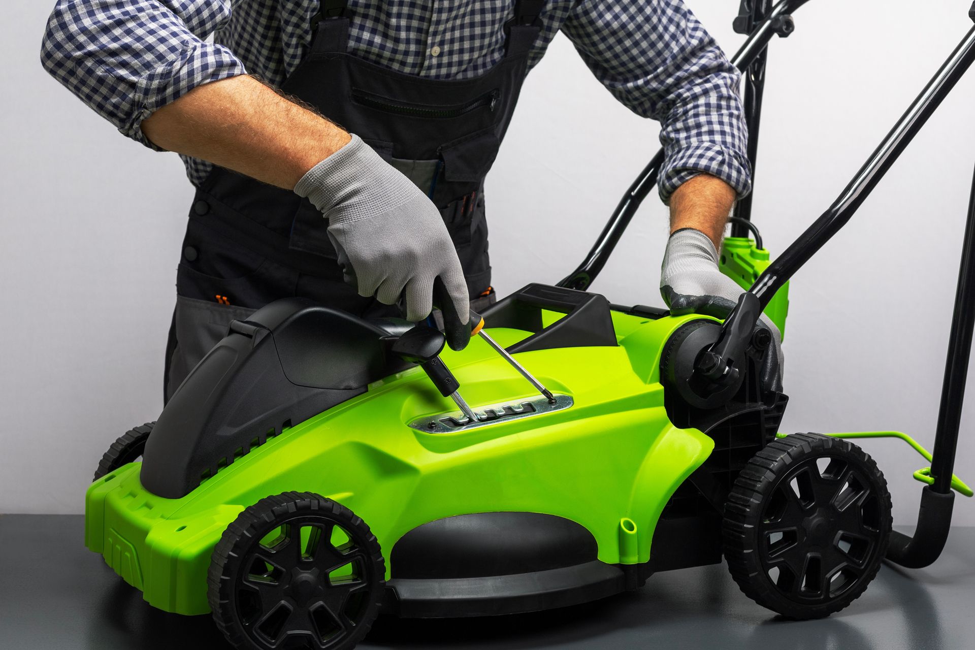Person in gray gloves repairing a bright green lawnmower with a screwdriver.