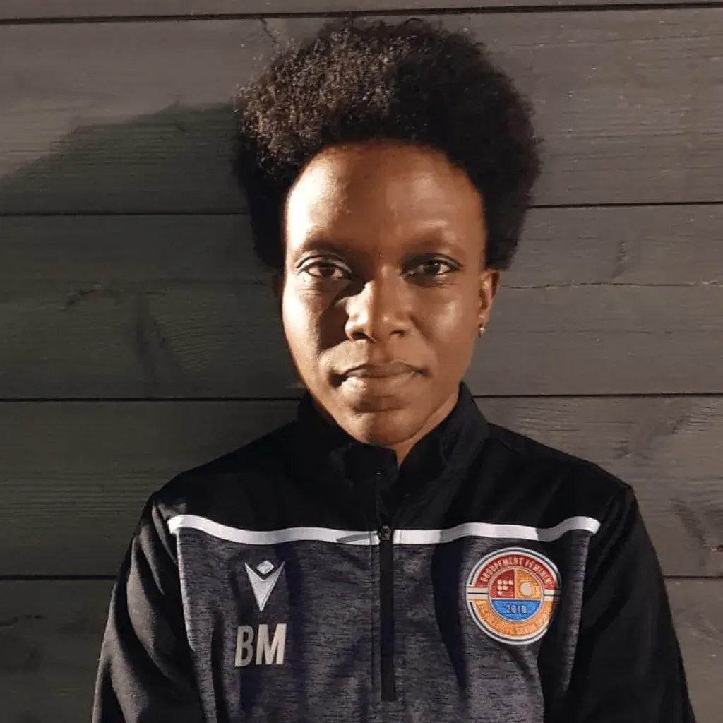 Une femme portant une veste noire avec le nom BM dessus se tient devant un mur en bois.