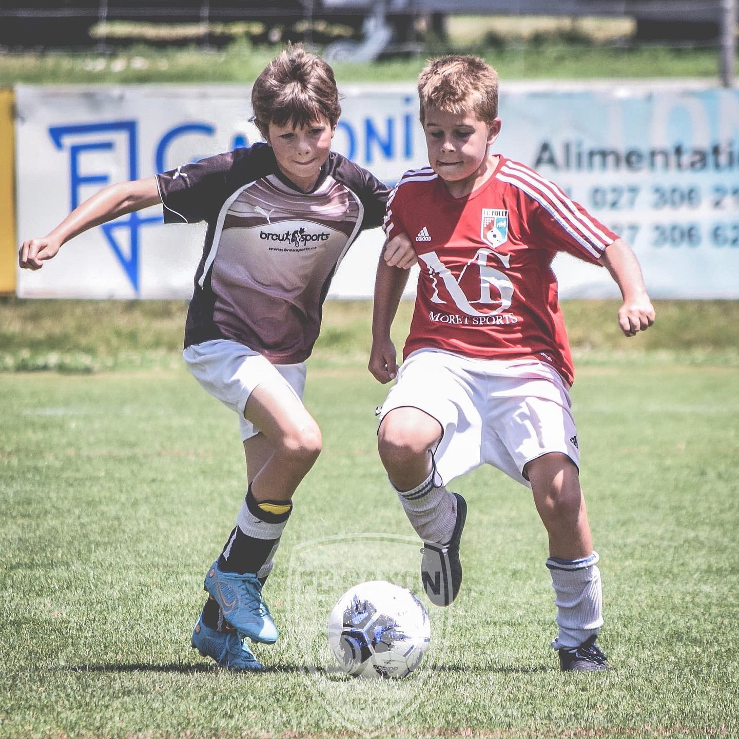 Deux jeunes garçons jouent au football sur un terrain avec un panneau en arrière-plan indiquant alimentati