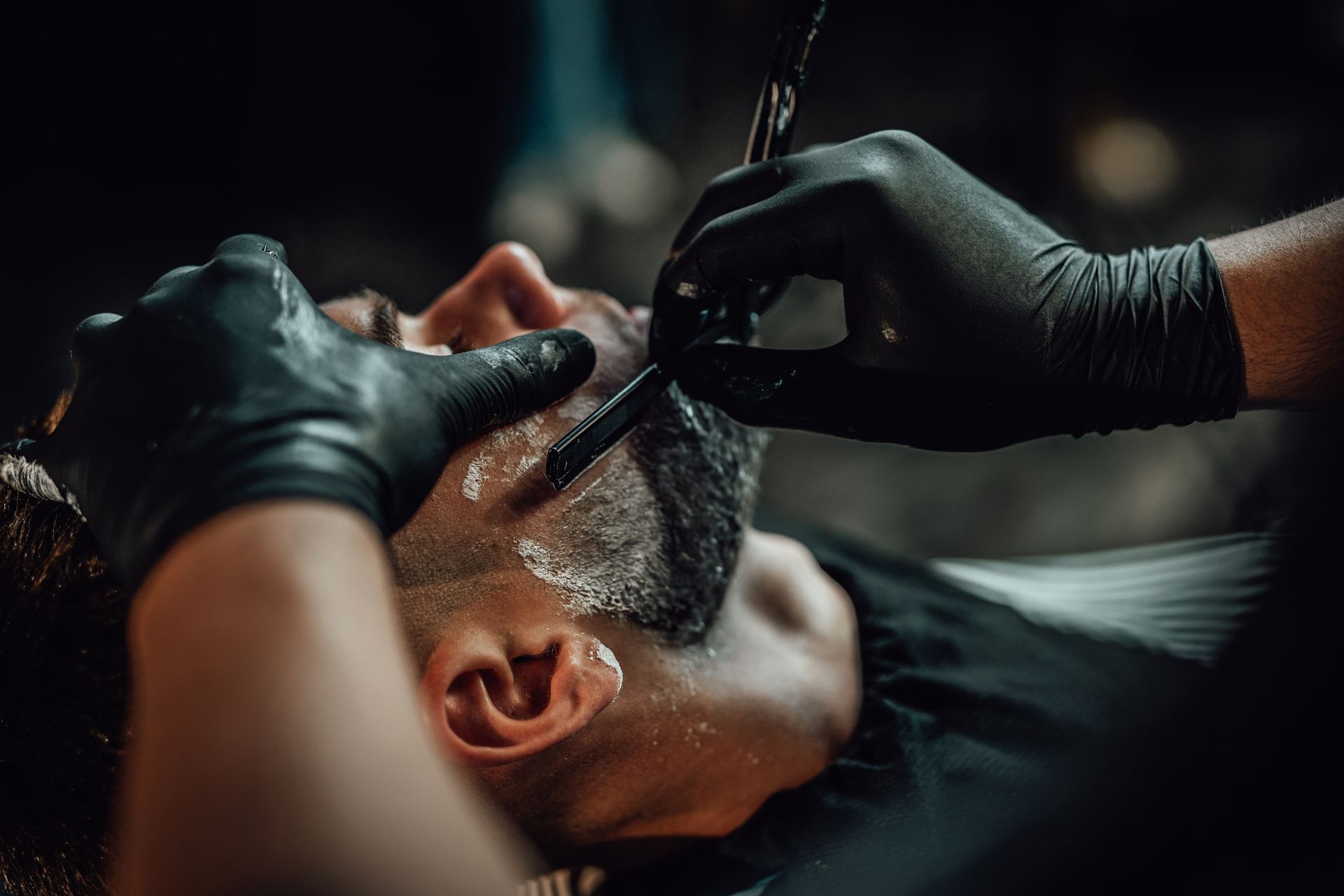 A man is getting his beard shaved by a barber wearing black gloves.