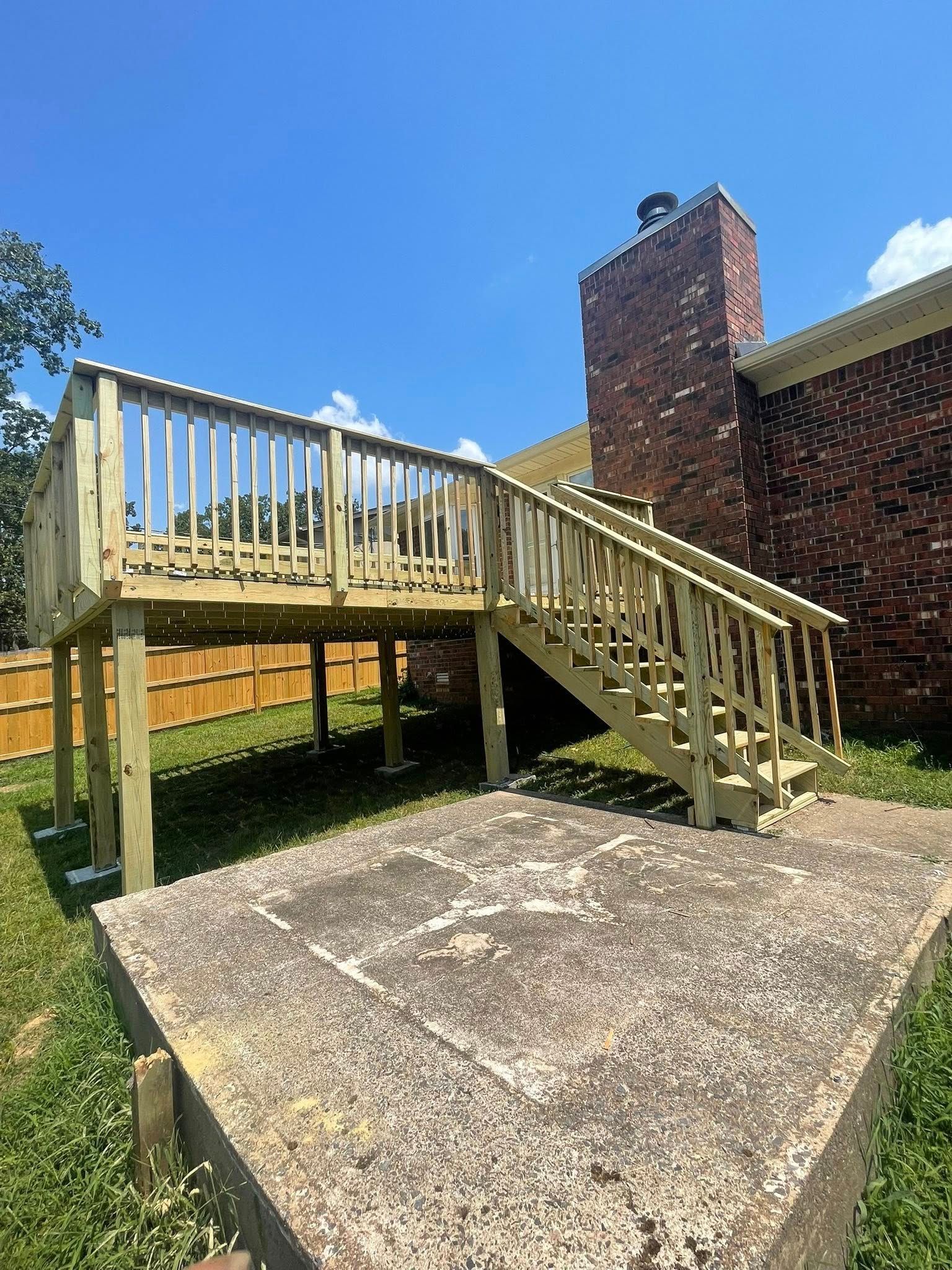 Wooden deck with stairs next to a brick chimney and concrete patio. Green grass and blue sky.