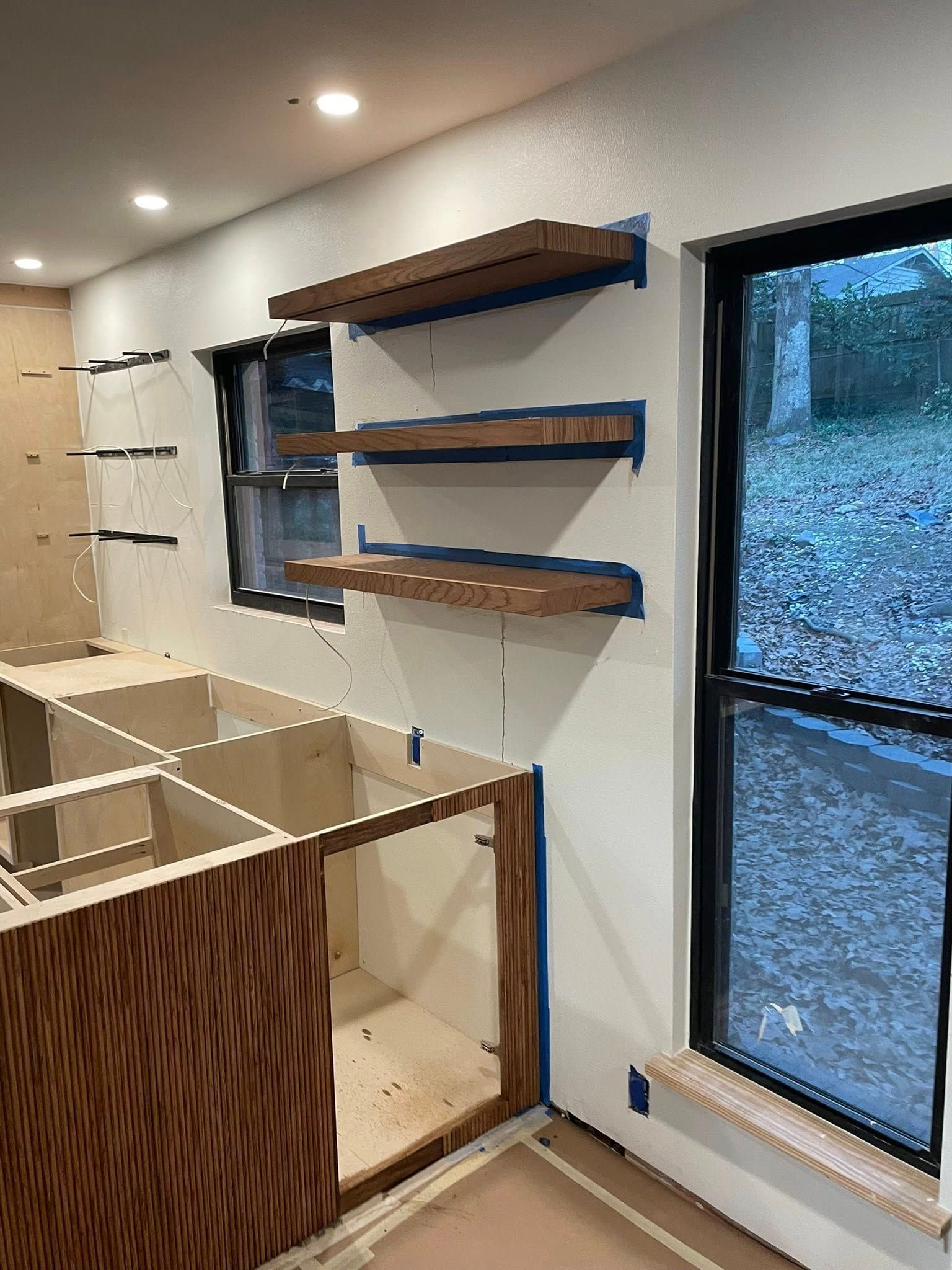 Kitchen under renovation with floating shelves, bare cabinets, and windows with black frames.