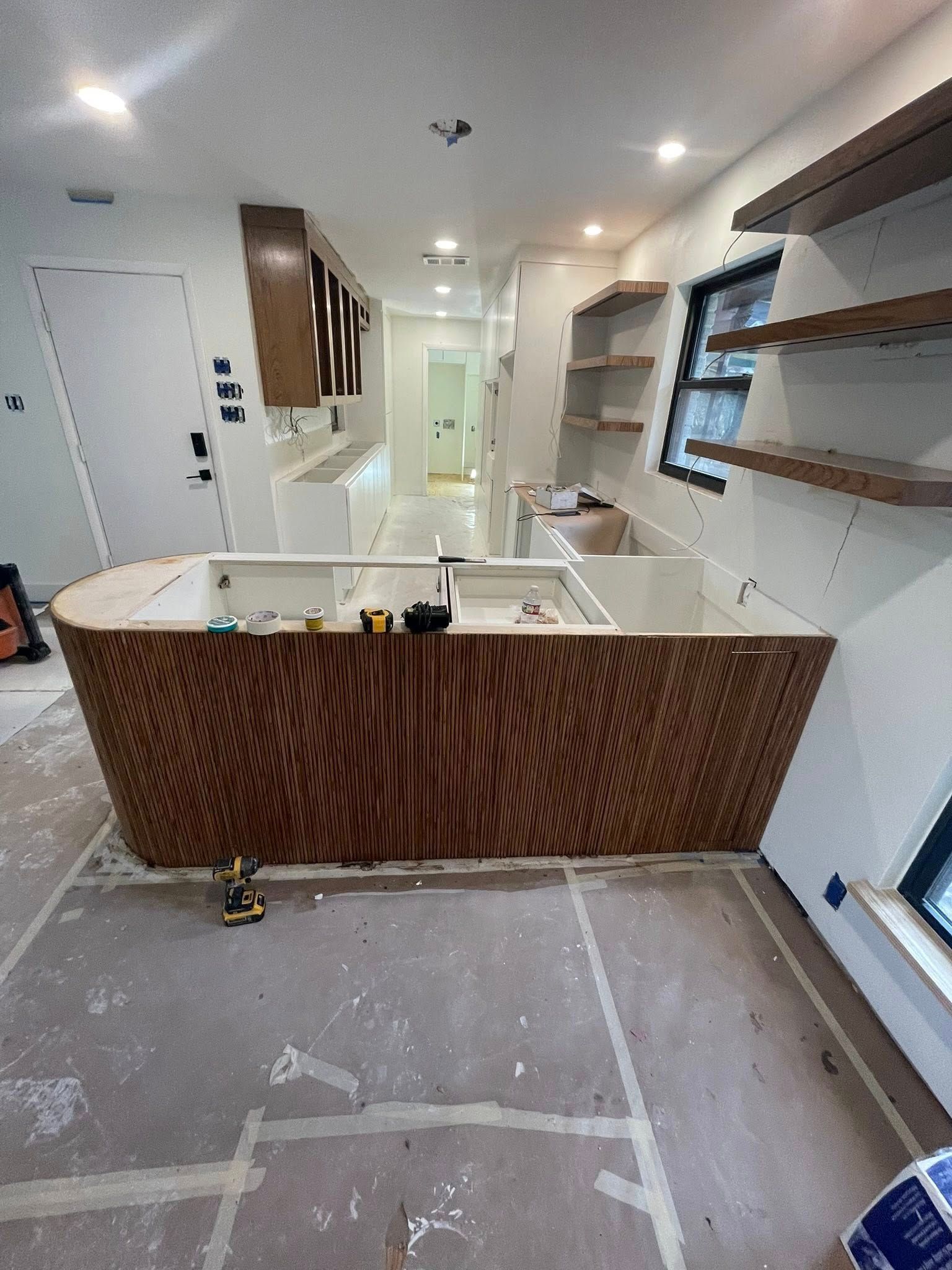 Kitchen under construction with curved wooden island, white cabinets, and floating shelves.